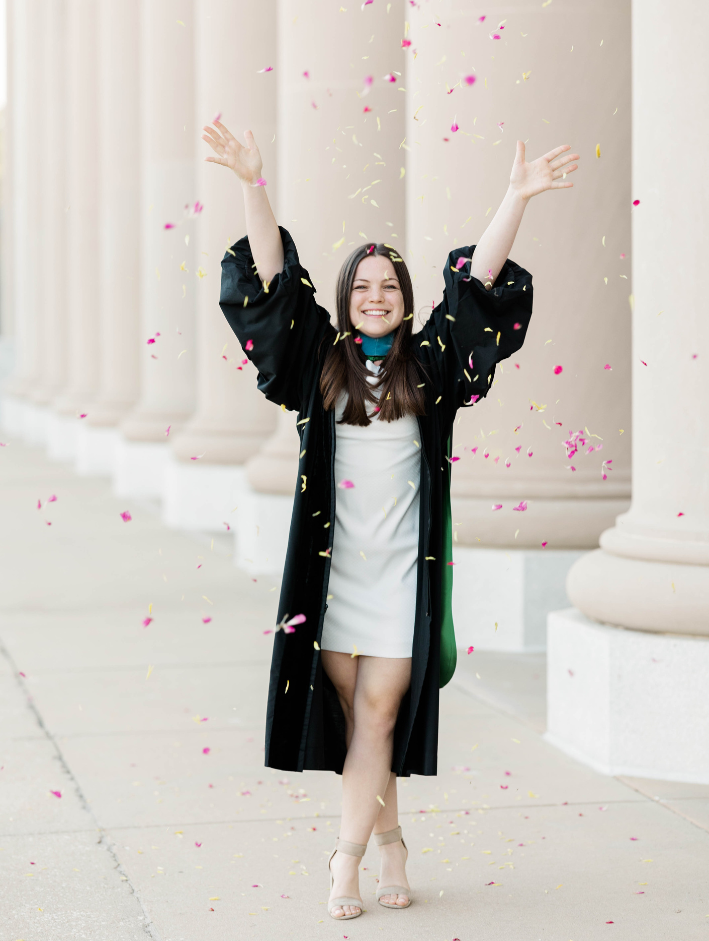 Jacksonville University doctoral graduate celebrating her spring graduation by tossing pink and yellow petals in the air during a joyful downtown Jacksonville graduation photo session in front of neoclassical columns.