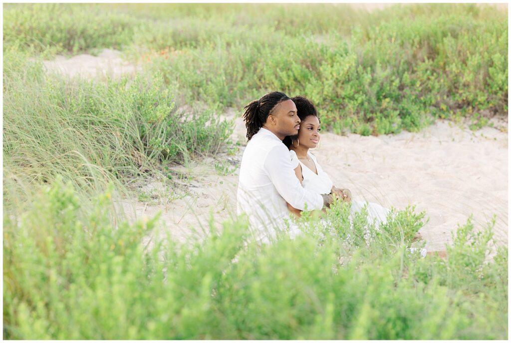 The couple sits closely in the dunes, surrounded by lush green beach grass and soft sand, gazing out quietly during their engagement session at Guana Beach Preserve in Ponte Vedra.