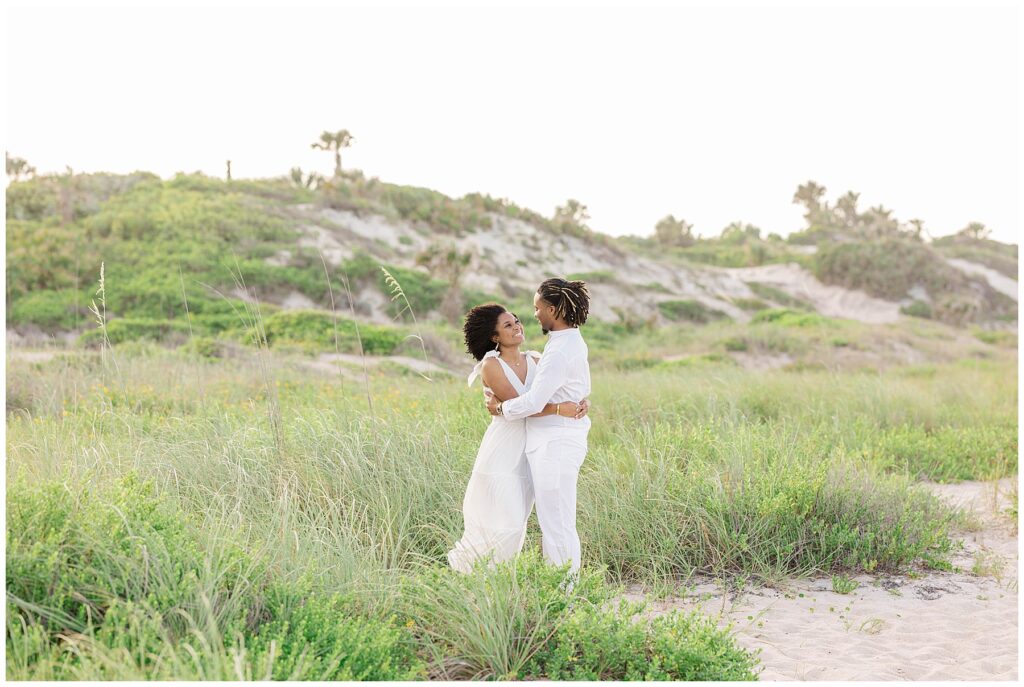 Romantic portrait of the couple embracing in tall beach grass with rolling dunes and wild greenery in the background.