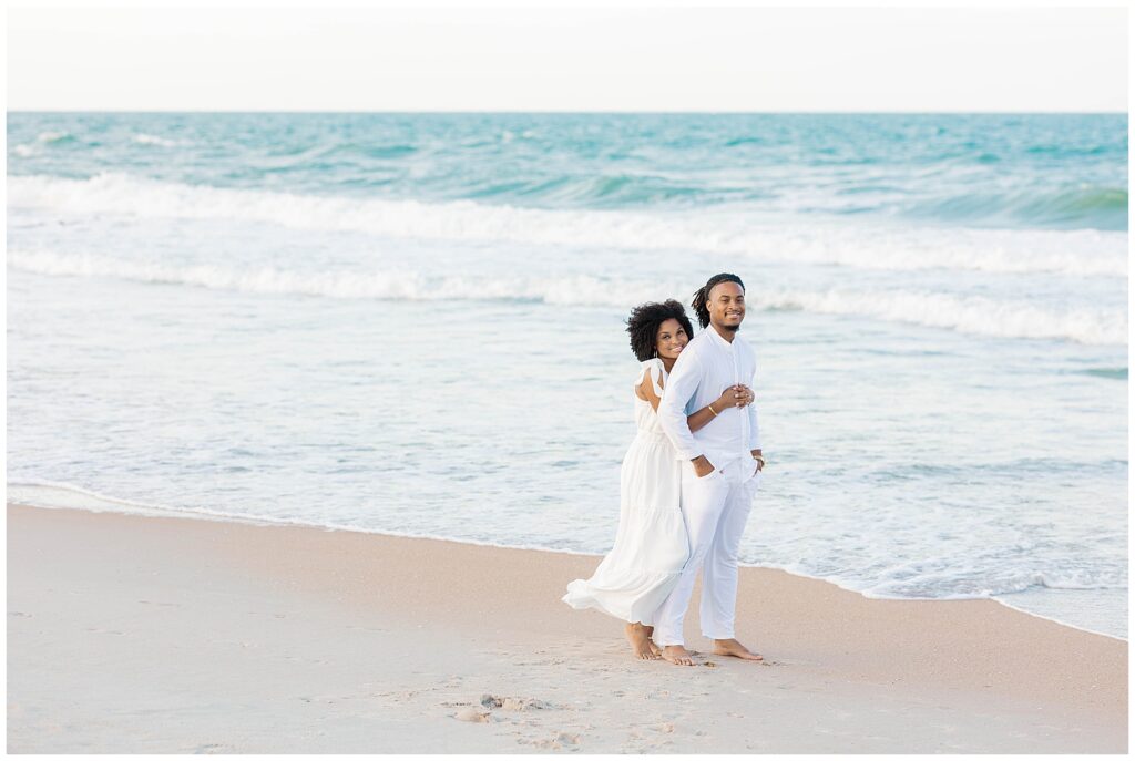 Bride-to-be leaning into her fiancé as they walk barefoot along the ocean’s edge in all-white outfits, captured during a peaceful and romantic engagement session at Guana Beach in Ponte Vedra.