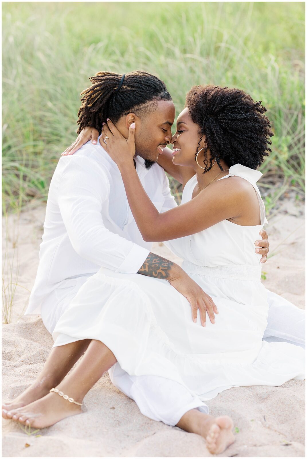  Intimate photo of the couple sitting barefoot in the sand, foreheads touching as they smile softly, surrounded by coastal grass during their beach engagement session.