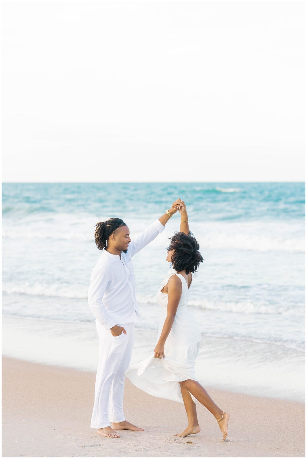 Groom-to-be twirling his fiancée barefoot on the shoreline, both dressed in white, with the ocean waves rolling in behind them during a romantic engagement session in Ponte Vedra.
