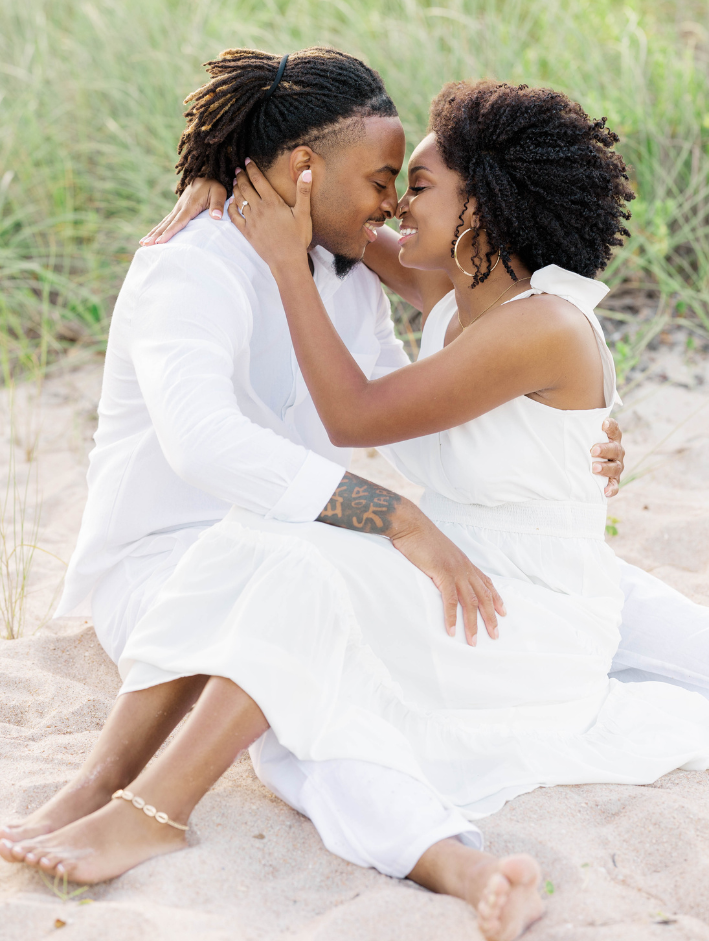 Couple sitting in the sand with foreheads touching, smiling softly during a quiet, romantic moment in the dunes