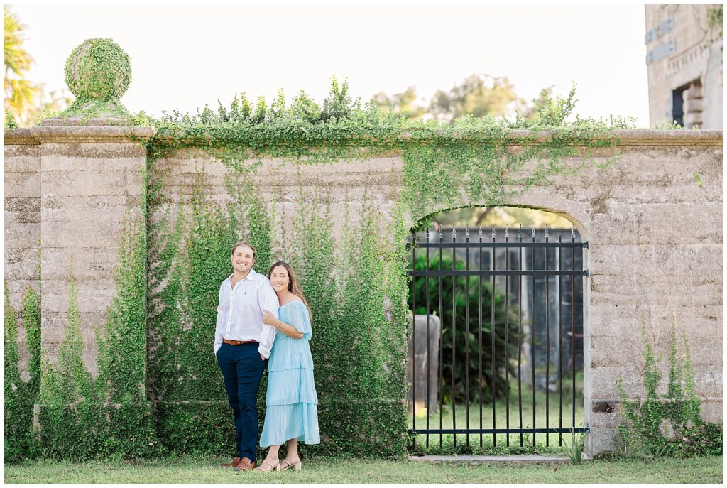 An engaged couple stands in front of an ivy-covered stone wall, smiling and holding hands on a sunny afternoon.