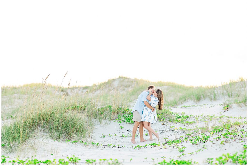 Romantic beach engagement photo of a couple sharing a kiss in the sand dunes surrounded by sea oats and greenery on Cumberland Island.