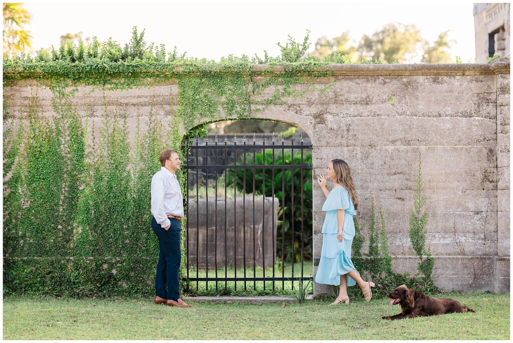 A woman playfully leans against a vine-covered wall as her partner smiles back from the opposite side, with their dog resting on the grass nearby.