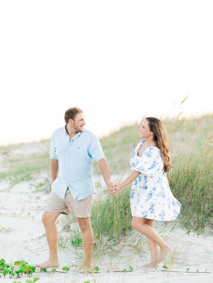 Couple walking hand-in-hand through the sand dunes during a romantic engagement session on Cumberland Island beach, smiling at each other barefoot in coastal grasses