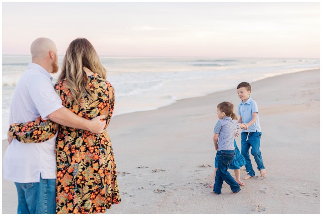 Parents watch their two young boys toss sand and play near the waterline while mom holds their daughter, all under a pink winter sunset at Guana Beach.
