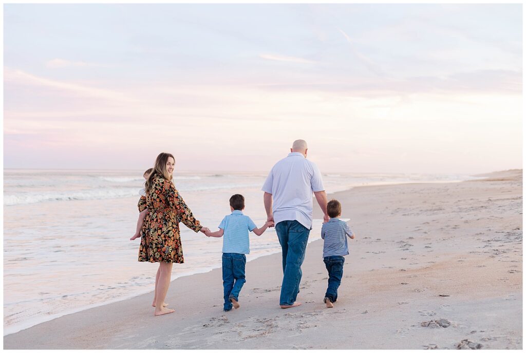 Family of five walking hand in hand along the shoreline at sunset, mom looking back and smiling while holding their daughter on Ponte Vedra Beach