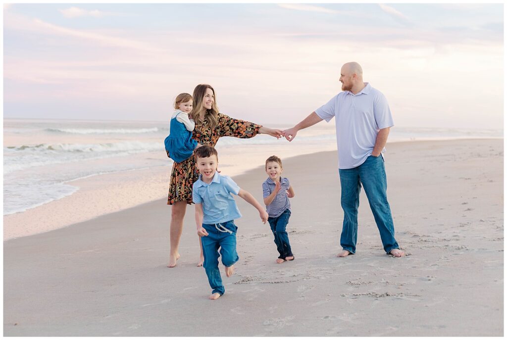 Two boys run joyfully toward the camera as mom and dad stretch arms across the sand, smiling with their daughter during a family session in Ponte Vedra Beach.