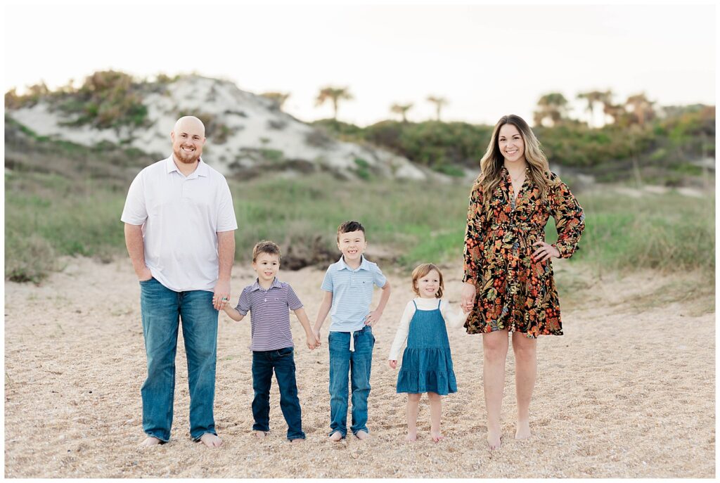 Family of five standing in a line on the beach in front of grassy dunes, each parent holding a child’s hand and smiling at the camera.
