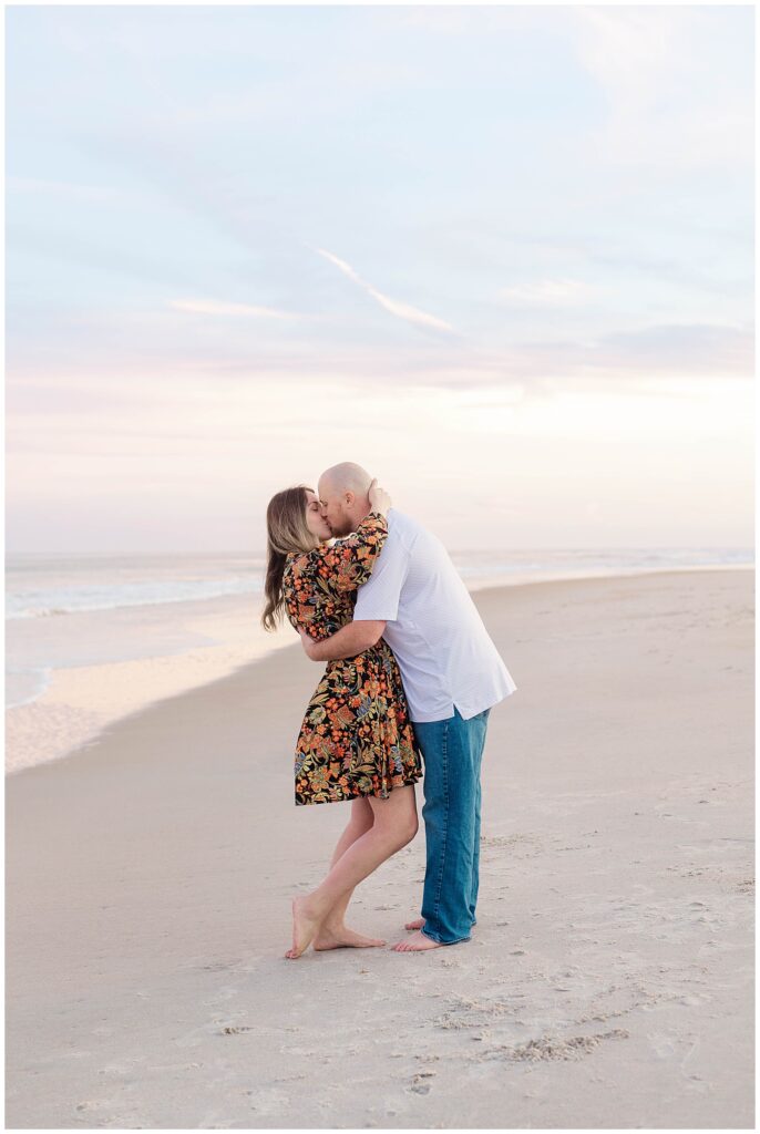 Mom and dad share a romantic kiss barefoot on the beach with a soft pink sunset behind them during their winter session at Guana Beach.