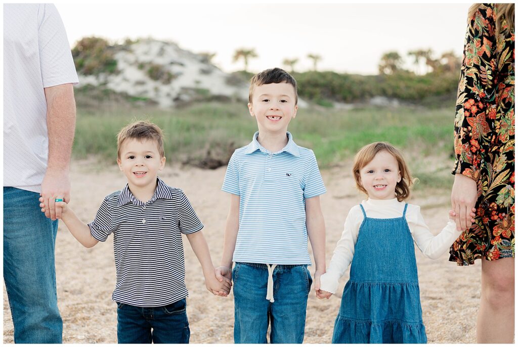 Three young siblings holding hands with their parents cropped at the edge of the frame, smiling during a family session in Ponte Vedra Beach.