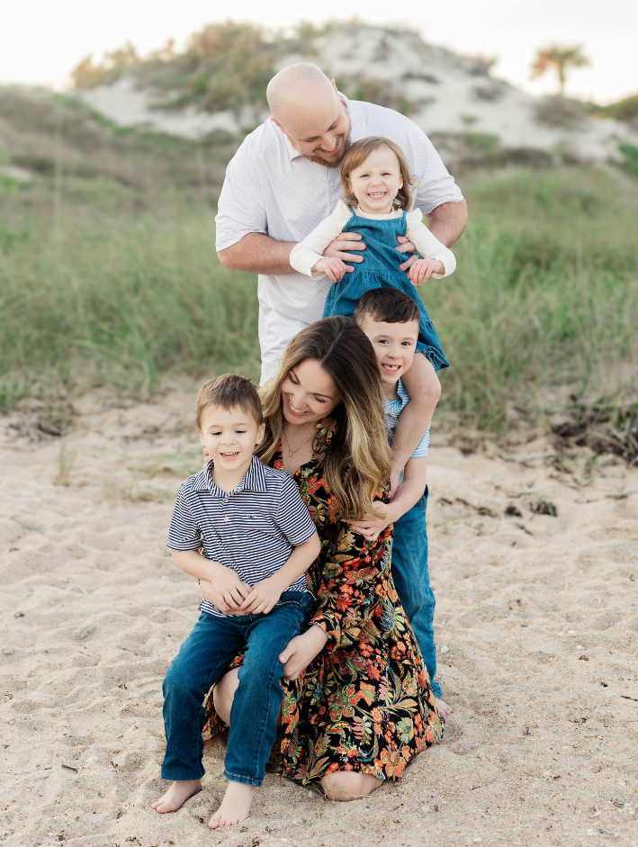 Family of five posing on the beach with father holding daughter on shoulders and mother sitting in the sand with two sons.