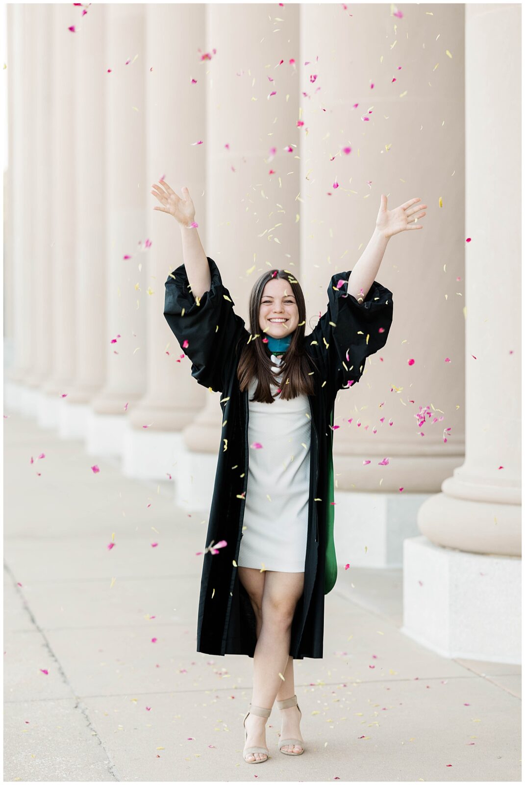 Jacksonville University doctorate graduate celebrating her spring graduation by tossing pink and yellow petals in the air during a downtown Jacksonville graduation photoshoot in front of historic columns.