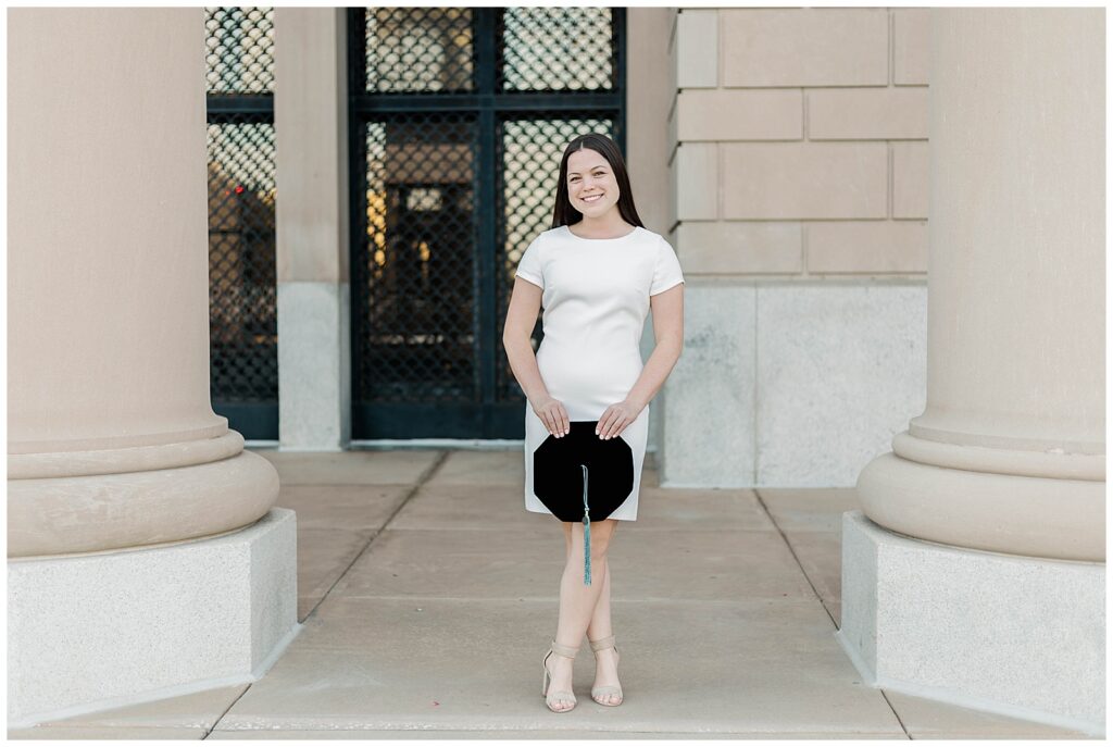 Jacksonville University doctoral graduate in a white dress posing between tall columns during her spring graduation photo session in downtown Jacksonville, holding her velvet tam and smiling warmly.