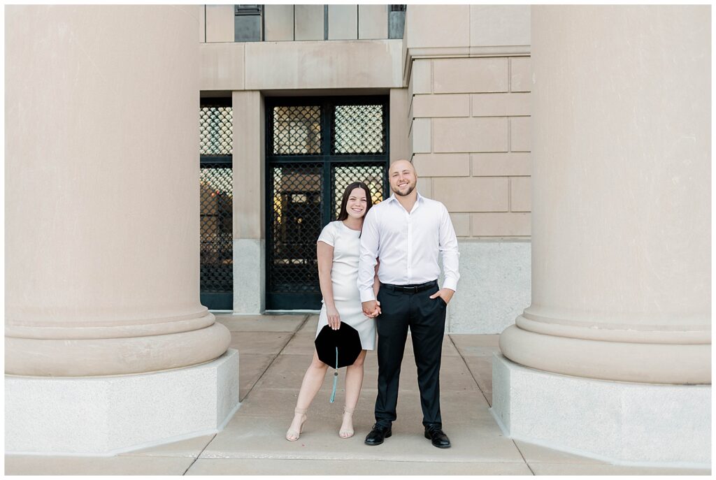 Jacksonville University doctoral graduate and her fiancé holding hands and smiling during their spring graduation photo session in downtown Jacksonville, posed between two large stone columns.