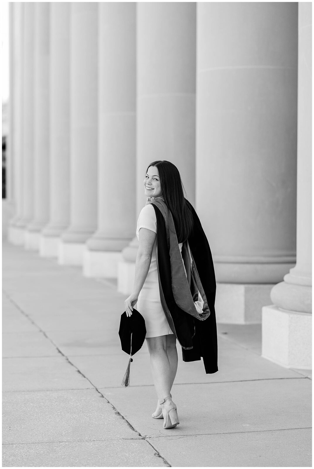 Black and white portrait of a Jacksonville University doctoral graduate walking away with a smile, holding her velvet tam and gown over her shoulder during a spring graduation photoshoot in downtown Jacksonville. 