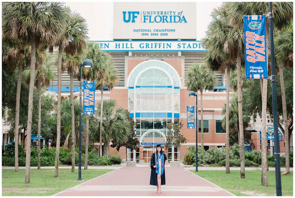 UF graduate standing centered in front of the entrance to Ben Hill Griffin Stadium, surrounded by palm trees and blue 'Gator Walk' banners leading up to the University of Florida sign.
