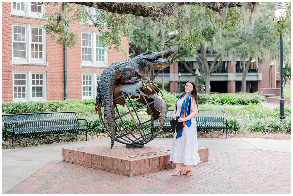 University of Florida graduate posing beside the alligator globe statue on campus, smiling in her white dress and blue graduation stole while holding her cap.