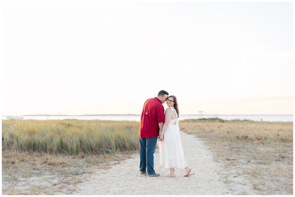 Engaged couple standing hand in hand on a sandy coastal path, with tall grasses and calm water in the distance during golden hour. 