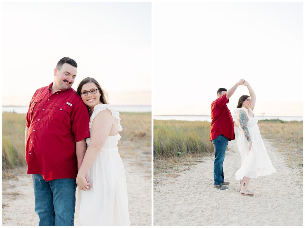 Engaged couple standing together and dancing along a sandy coastal trail, smiling and holding hands with tall grass and water in the distance.