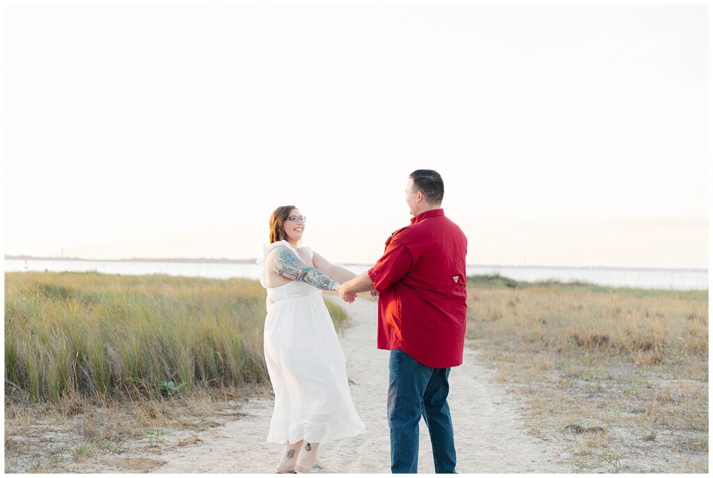 Engaged couple holding hands and smiling while standing on a sandy coastal path with tall grass and water behind them.