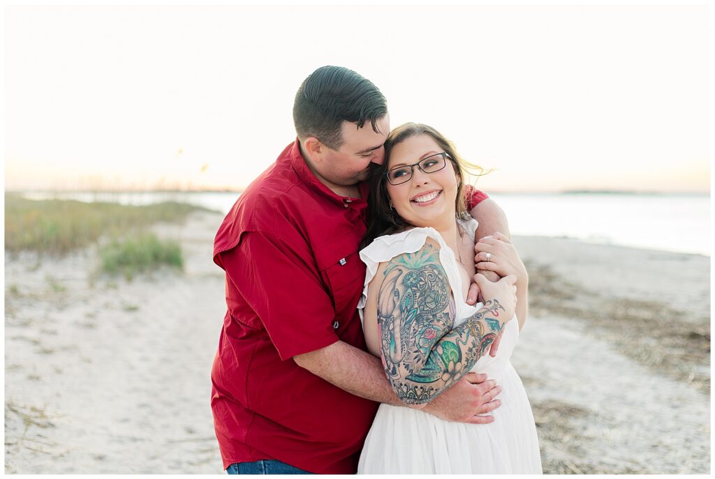 Wrapped in each other’s arms near the beach, one partner smiles toward the camera while the other leans in close during a Fort Clinch engagement session in Fernandina Beach.