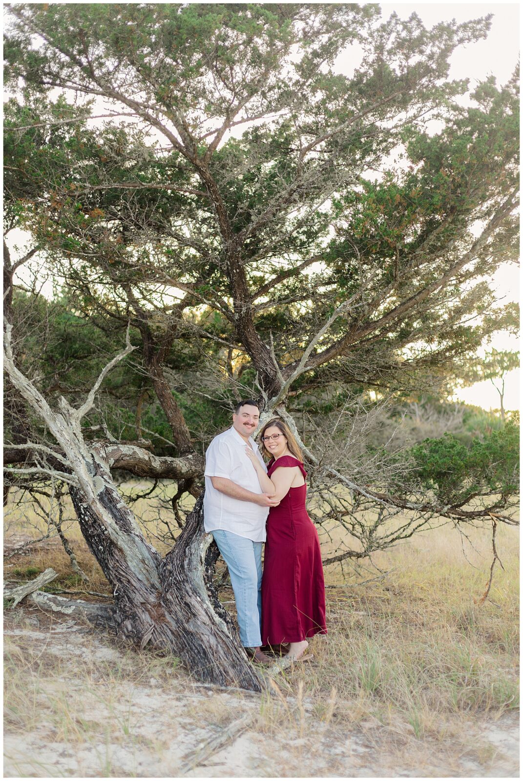 The couple stands together beneath a sprawling coastal tree, framed by tall grass and soft evening light.