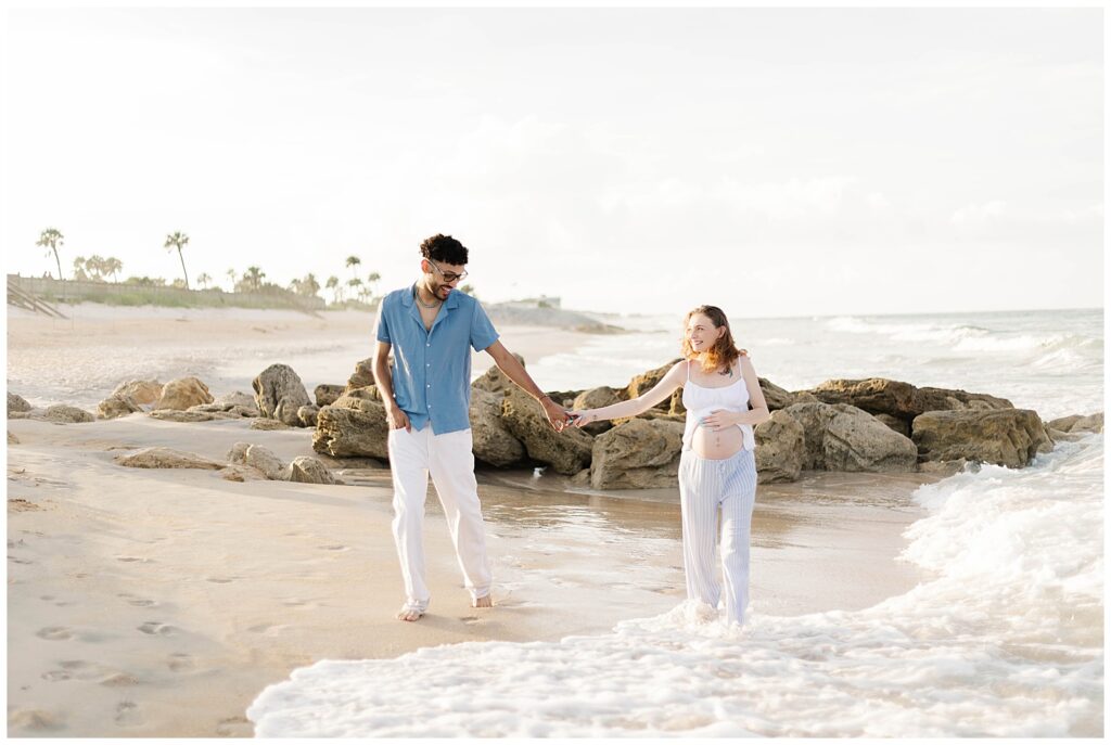 Expecting parents holding hands while walking through shallow ocean water during golden hour