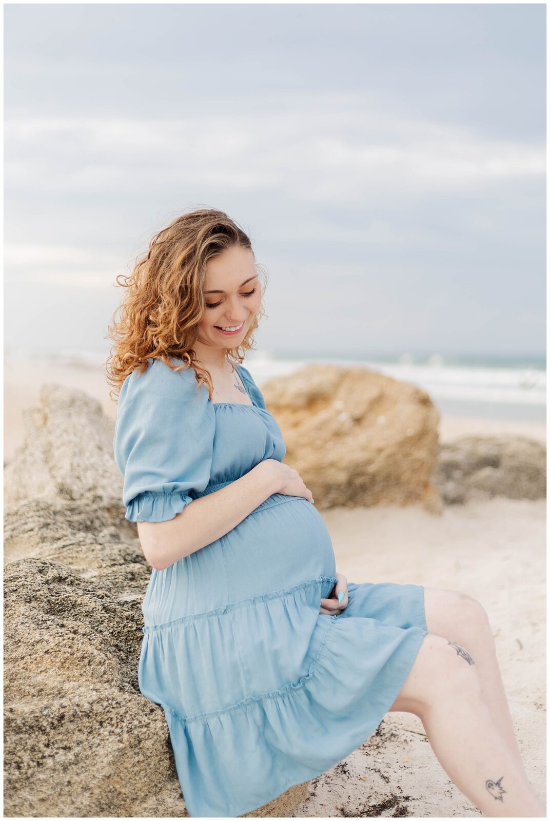 Pregnant mother seated on a rock at the beach, gently holding her baby bump and smiling softly