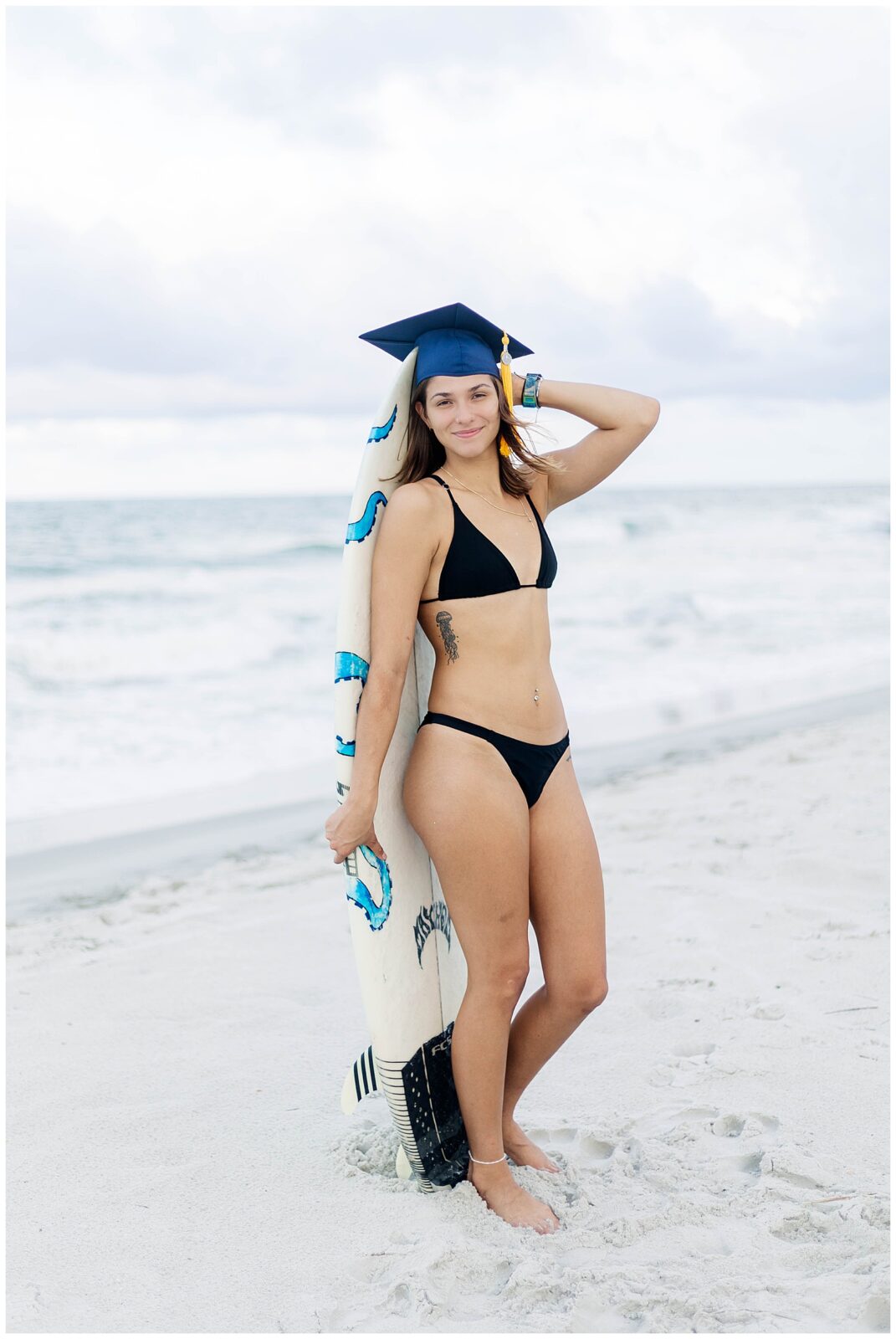 UNF graduate holding her surfboard and wearing her graduation cap while standing along the shoreline