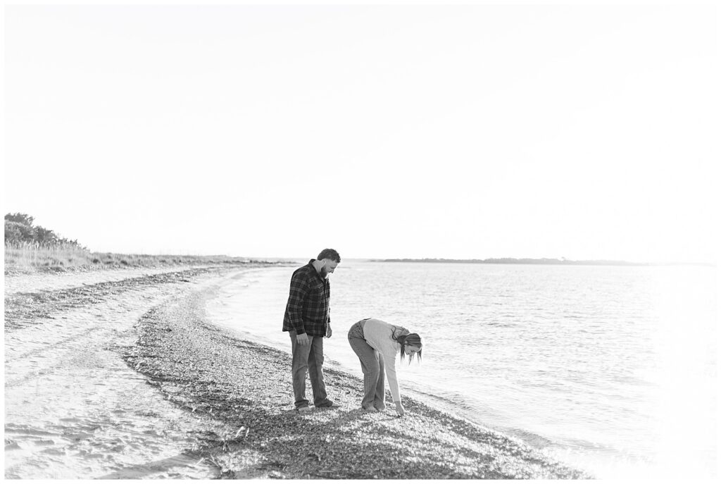 Fort Clinch State Park Engagement Black and white Fort Clinch engagement photo of couple walking along the shoreline as she bends to look at seashells and he watches nearby