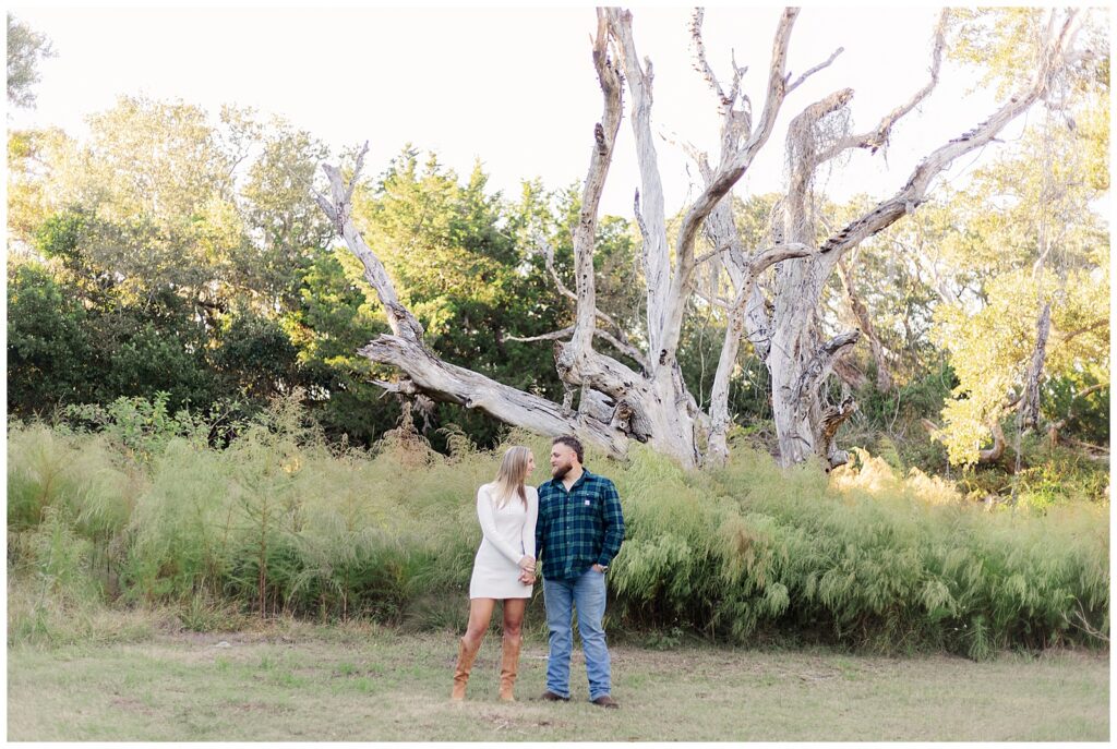 Fort Clinch Engagement Photos Engaged couple standing together in front of a striking driftwood tree surrounded by tall greenery at Fort Clinch State Park.