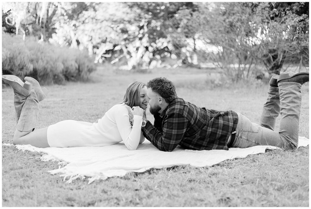Black and white portrait of the couple smiling at each other while lying on a blanket in a grassy field.