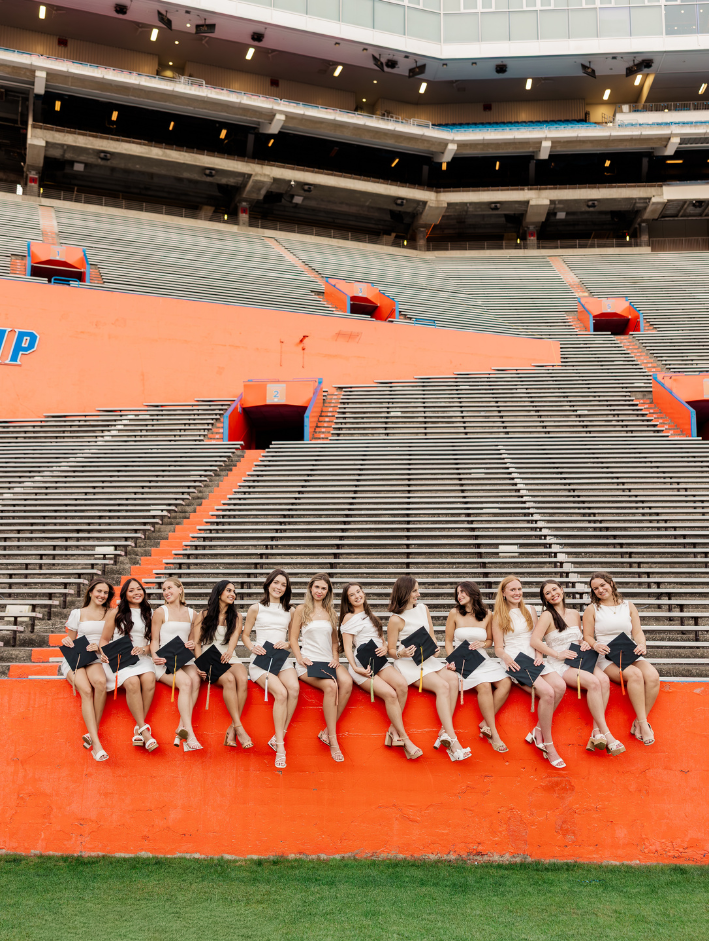 UF Pi Beta Phi grads sitting on the football field wall at the University of Florida holding graduation caps