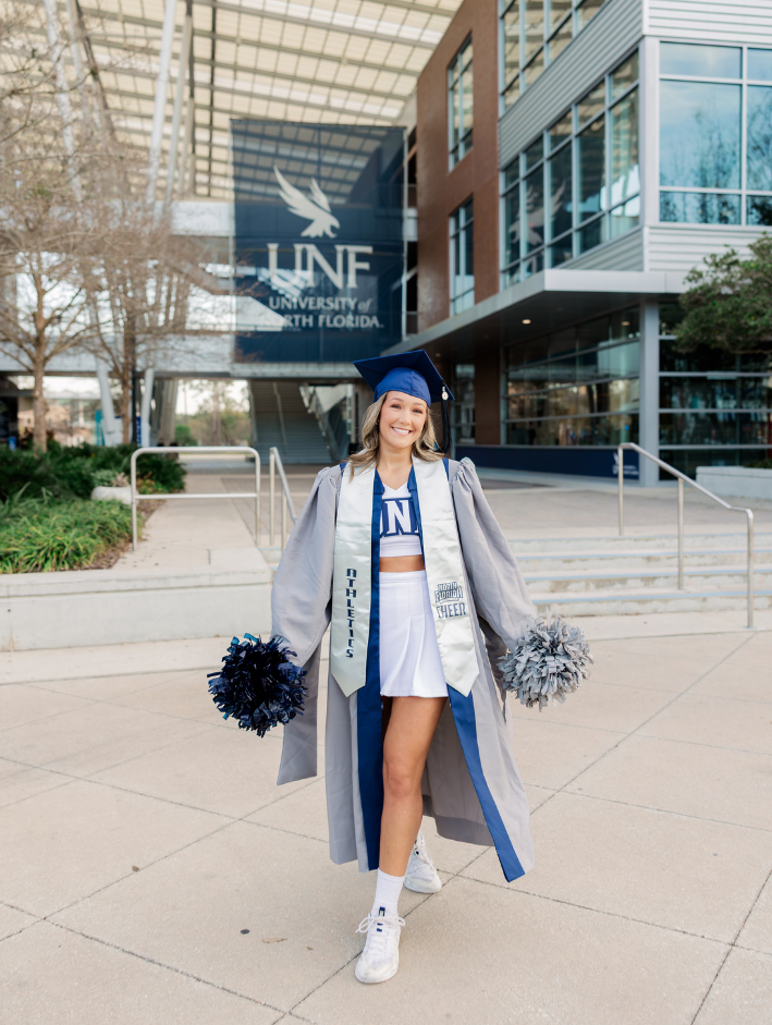 University of North Florida master’s graduation photos of UNF cheerleader in cap and gown holding pom poms on campus in Jacksonville Florida